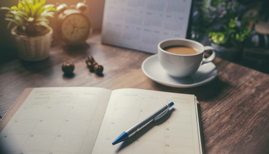 A planner on a desk with a pen, surrounded by a coffee cup and a small plant)